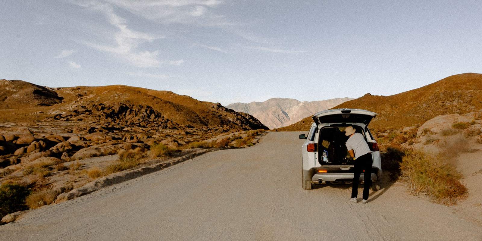 Pessoa de chapéu organiza a bagagem no porta-malas aberto de um carro branco parado numa estrada de terra entre morros secos